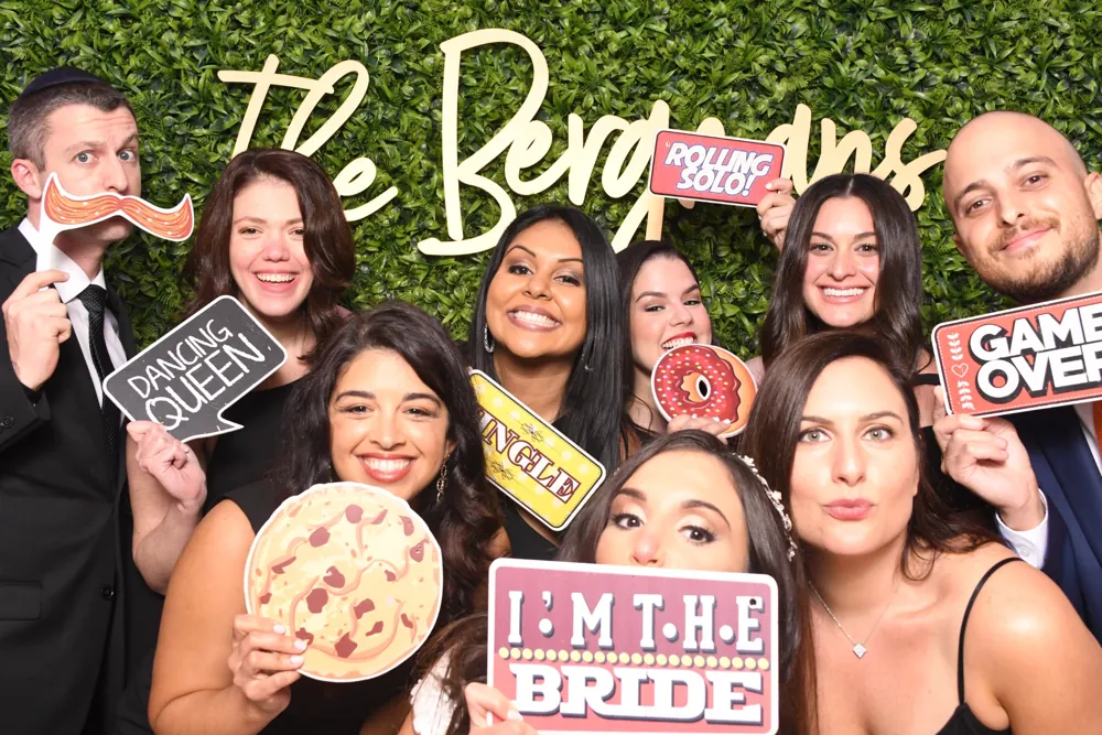 Wedding guests posing with playful props in front of a greenery backdrop